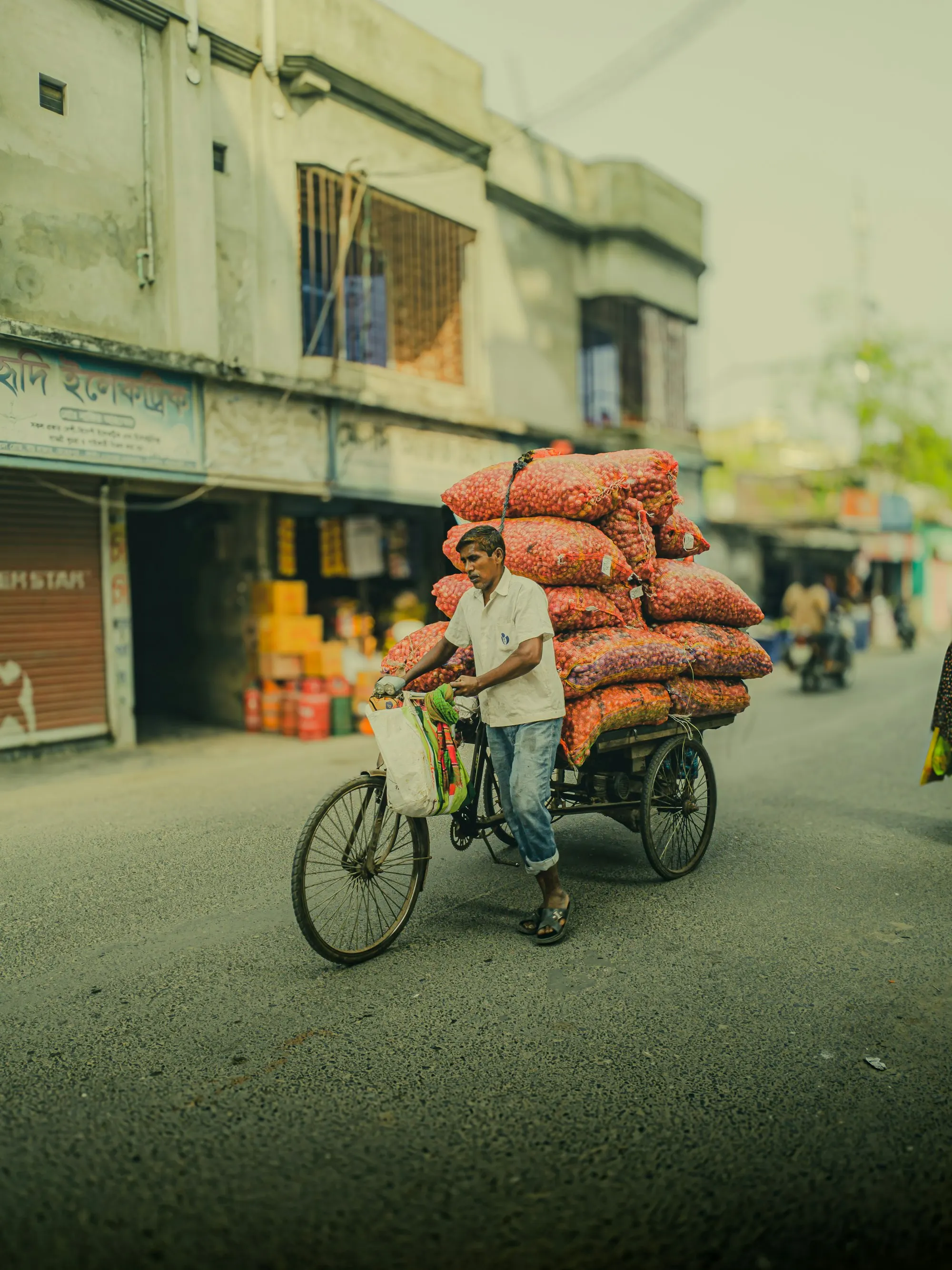 Photo: Ashikul Islam Anik, Unsplash A man pedals a three-wheeled van loaded high with orange sacks down a quiet street lined with shopfronts.